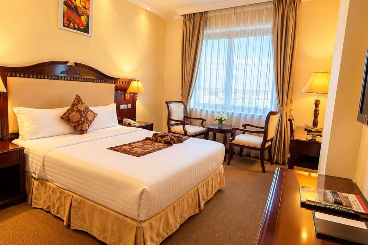 A hotel room with a large bed, two bedside tables, a desk, a chair, and a window with curtains.