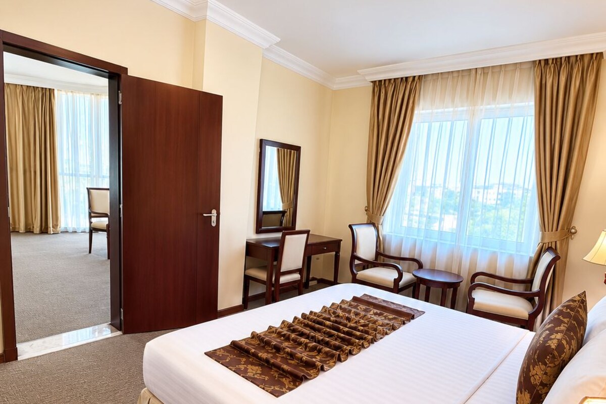 A hotel room with a large bed, a desk, two chairs, a mirror, and a window with curtains.