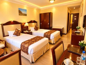 A hotel room with two beds, a desk, a chair, a wardrobe, and a bathroom visible in the background.