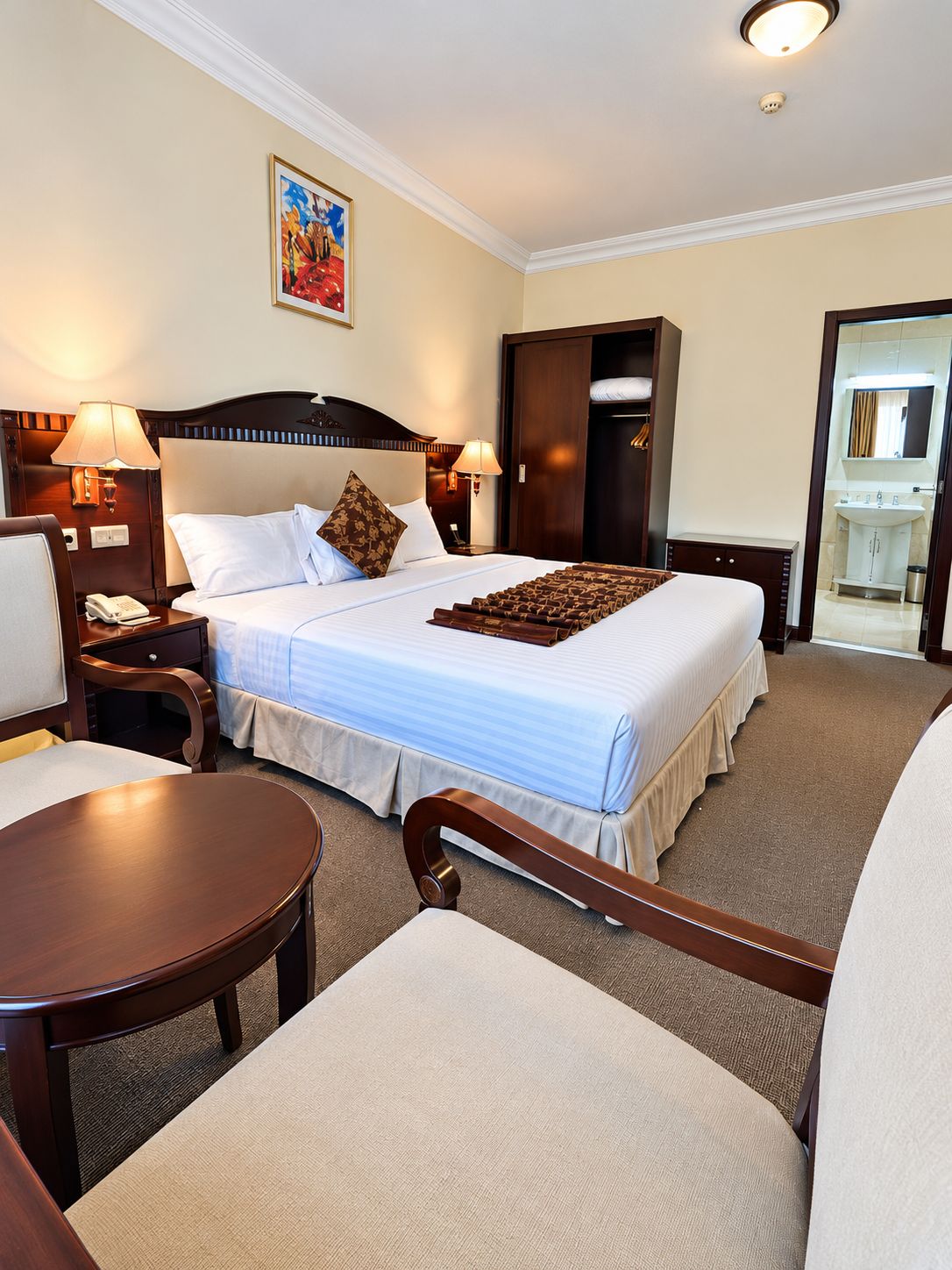 A hotel room with a large bed, two bedside tables with lamps, a chair, a desk, and a bathroom visible through an open door.