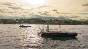 A boat named 'Reef Rider' on calm water with a backdrop of a cloudy sky and a distant shoreline with buildings and trees. - Liquid Dive Resort (Dauin)