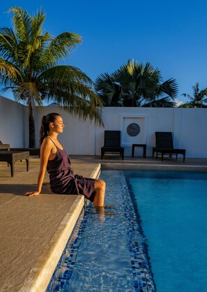 Una mujer sentada junto a una piscina con sillas de descanso y palmeras al fondo.