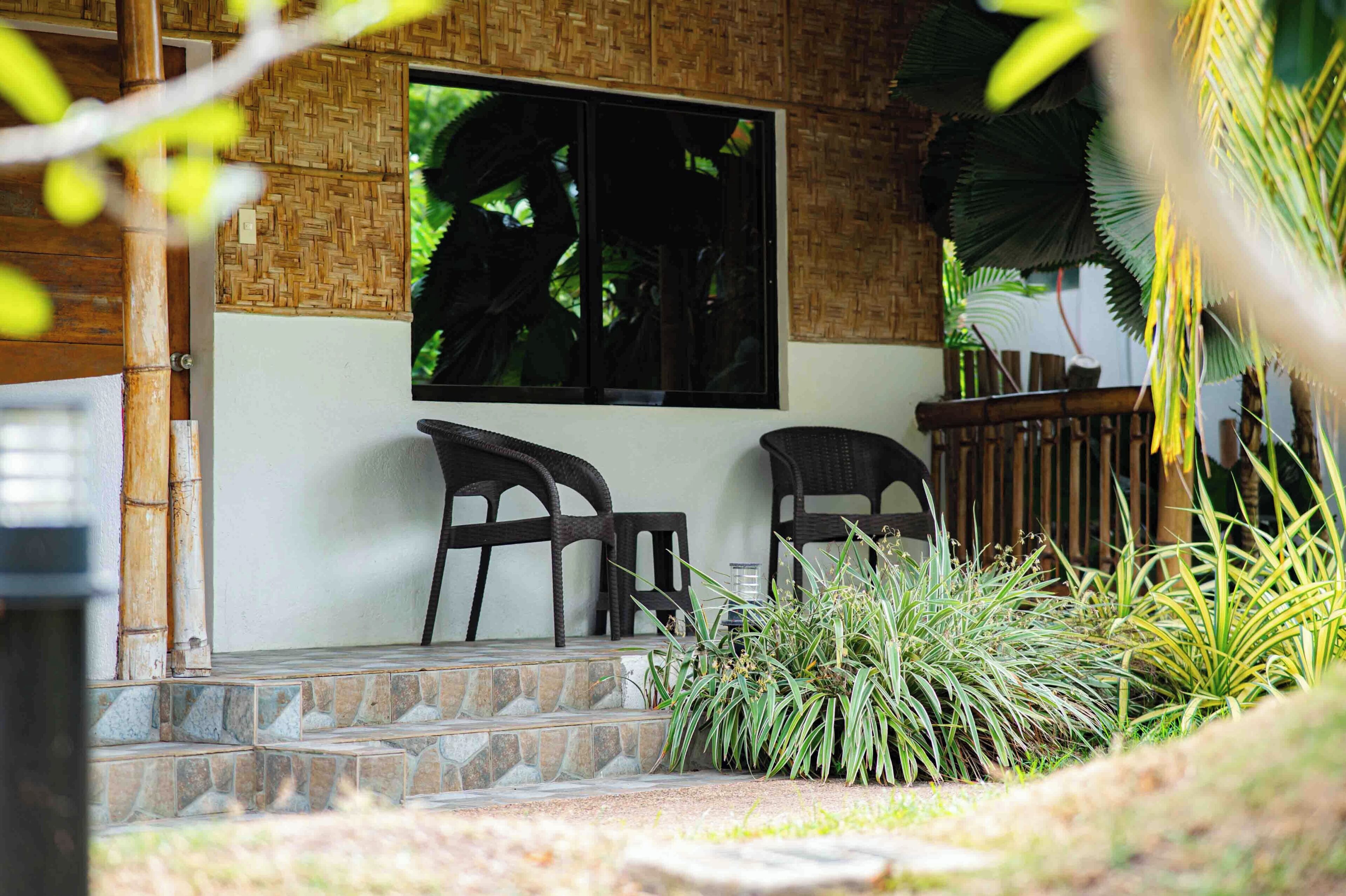 A covered outdoor seating area with two chairs and a table, surrounded by plants and a stone staircase.