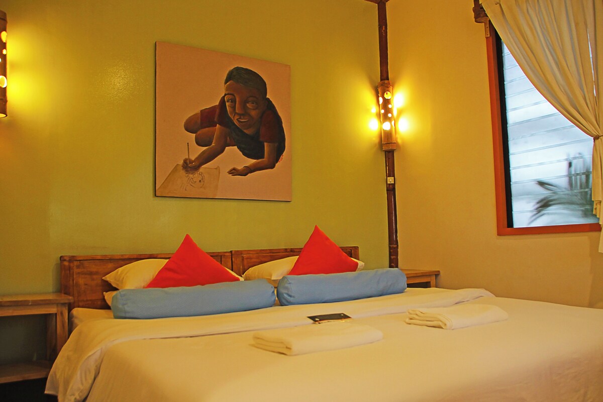 Una habitación de hotel con cama, almohadas, un cuadro en la pared y una ventana con cortinas.