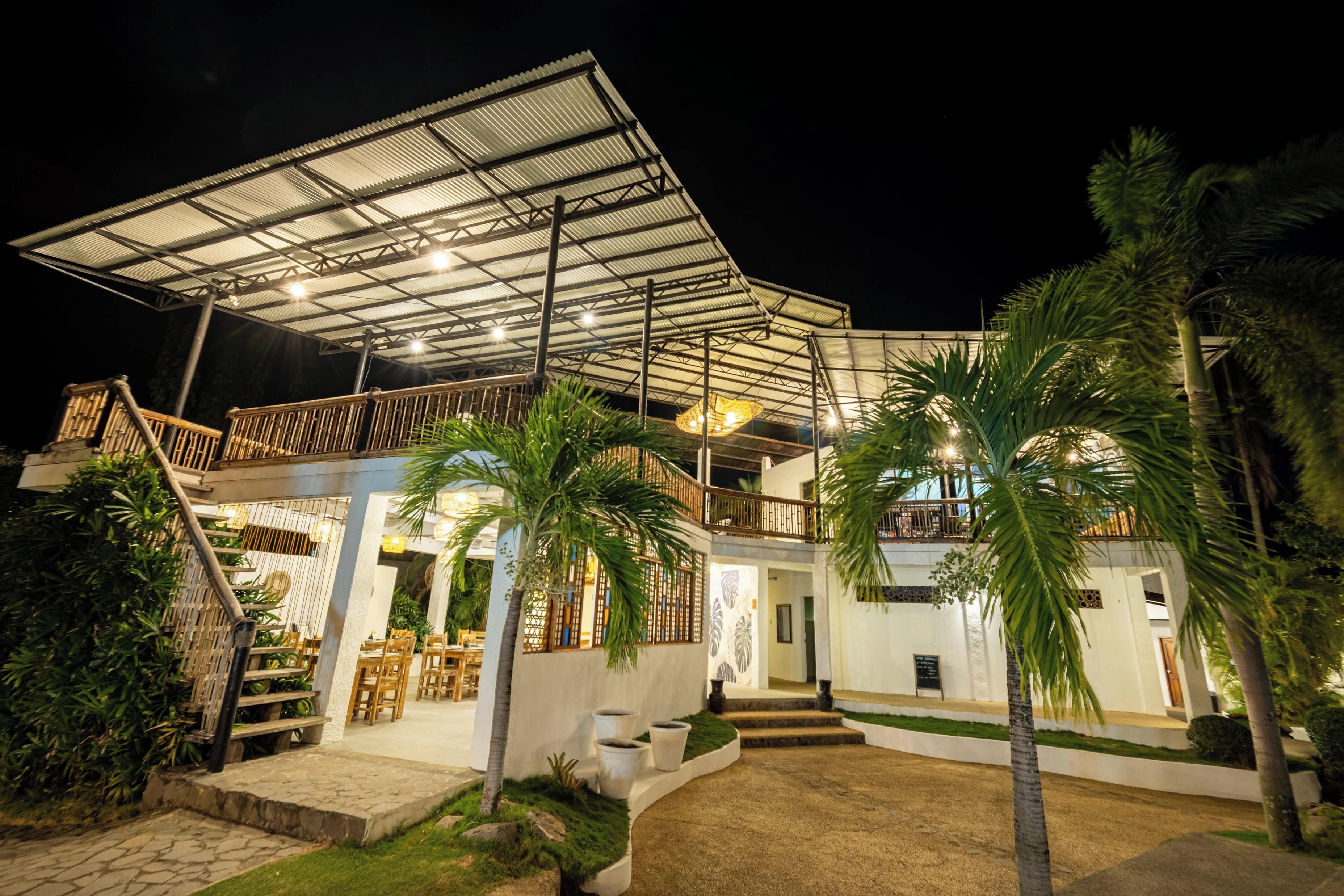 Un edificio de dos pisos con un área de estar al aire libre techada, palmeras y una escalera que conduce al nivel superior.
