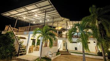 Un edificio de dos pisos con un área de estar al aire libre techada, palmeras y una escalera que conduce al nivel superior.