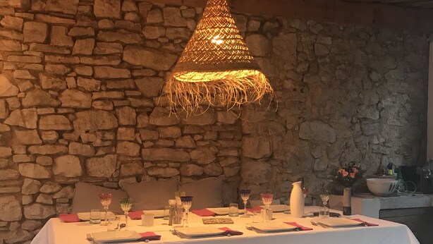 A dining table set for two with a stone wall backdrop, a woven light fixture above, and a bench with pillows in front. - Charming village house in Aix en Provence (Aix en Provence)