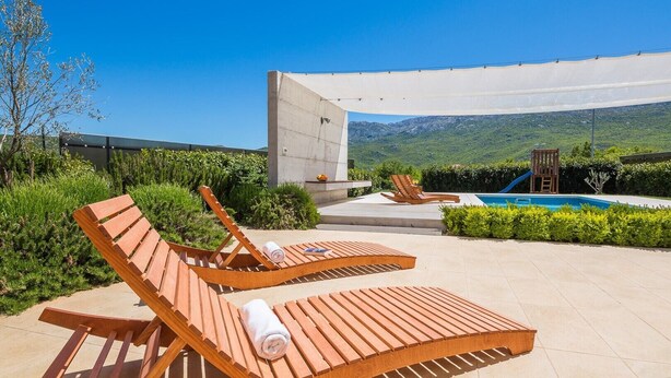 A poolside area with wooden loungers, a pool, and a view of mountains.