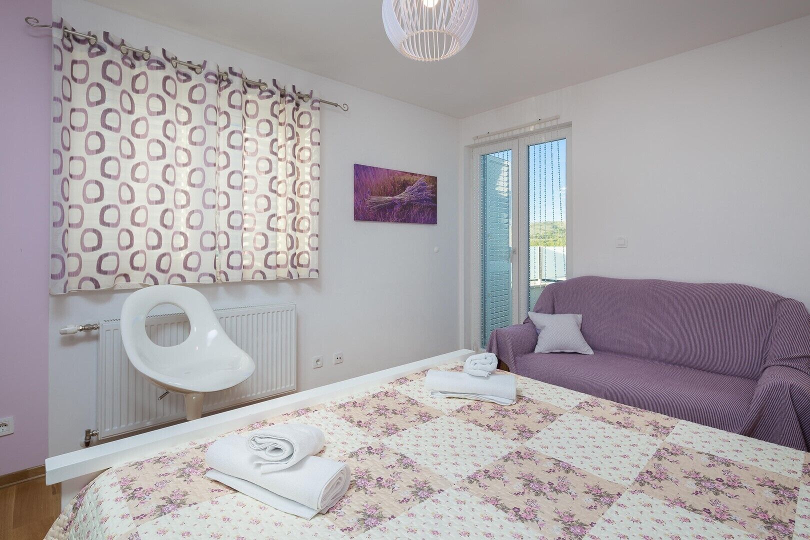 A bedroom with a bed, a purple sofa, a radiator, a window with curtains, and a painting on the wall.
