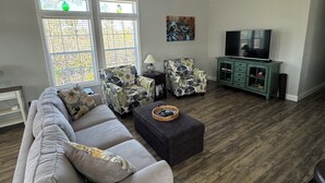 Un salon avec un canapé gris, deux fauteuils à motifs, une table basse, un téléviseur posé sur un meuble vert et un ventilateur de plafond.