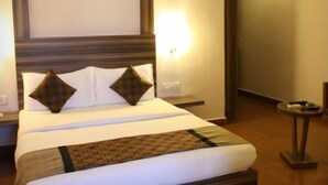Quarto de hotel com uma cama grande, duas almofadas, uma mesinha de cabeceira com abajur e uma pequena mesa com um controle remoto.