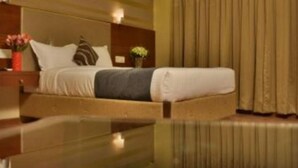 Una habitación de hotel con una cama, una mesita de noche, un florero con flores y una ventana con cortinas.