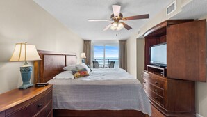 A hotel room with a bed, a TV, a ceiling fan, a nightstand, and a view of the outdoors.