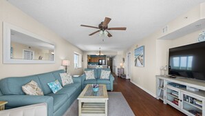 A living room with a teal sofa, a wooden coffee table, a ceiling fan, a TV, and a bookshelf.