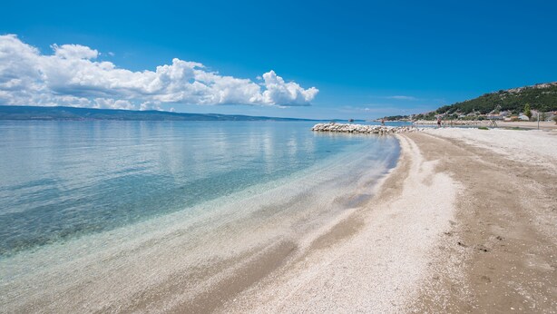 A beach with clear water, a rocky breakwater, and a distant shoreline with buildings. - Hotel Nestos (Dugi Rat)