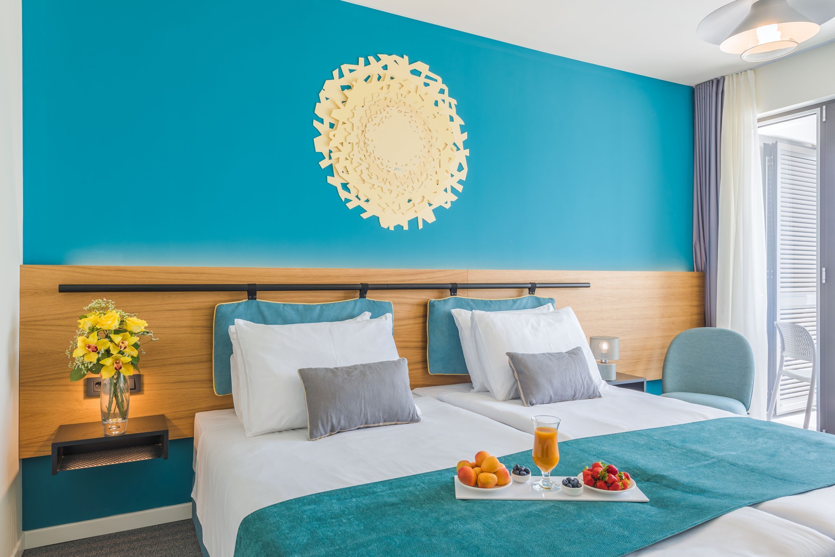 Ein Hotelzimmer mit einer türkisblauen Akzentwand, einem Wanddekor, einem Bett mit weißen Leinen, einem Nachttisch mit Lampe, einem Sessel und einer Ablage mit Obst und Saft.