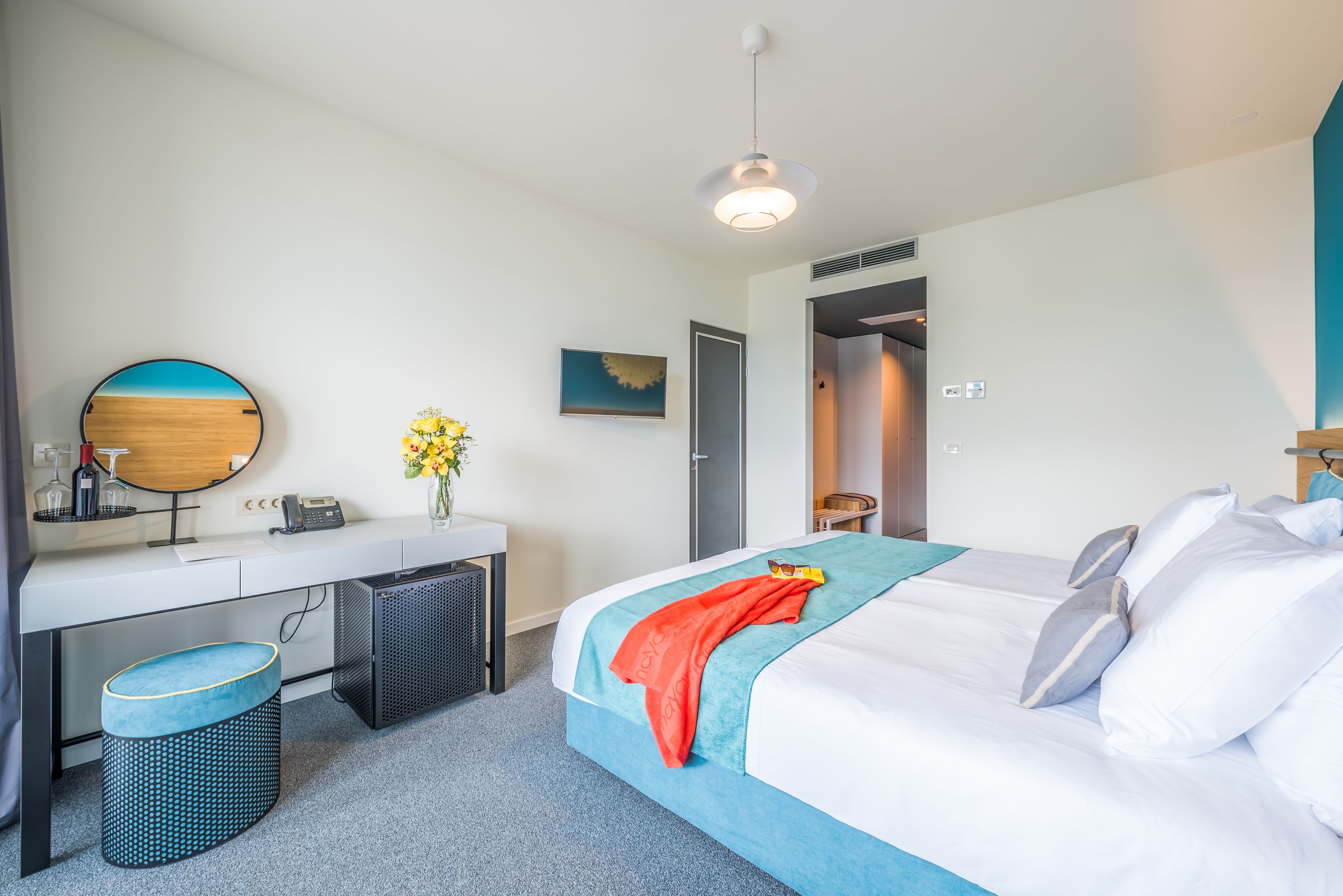 A modern hotel room with a bed, a desk, a chair, a small stool, a mirror, and a door leading to another room.