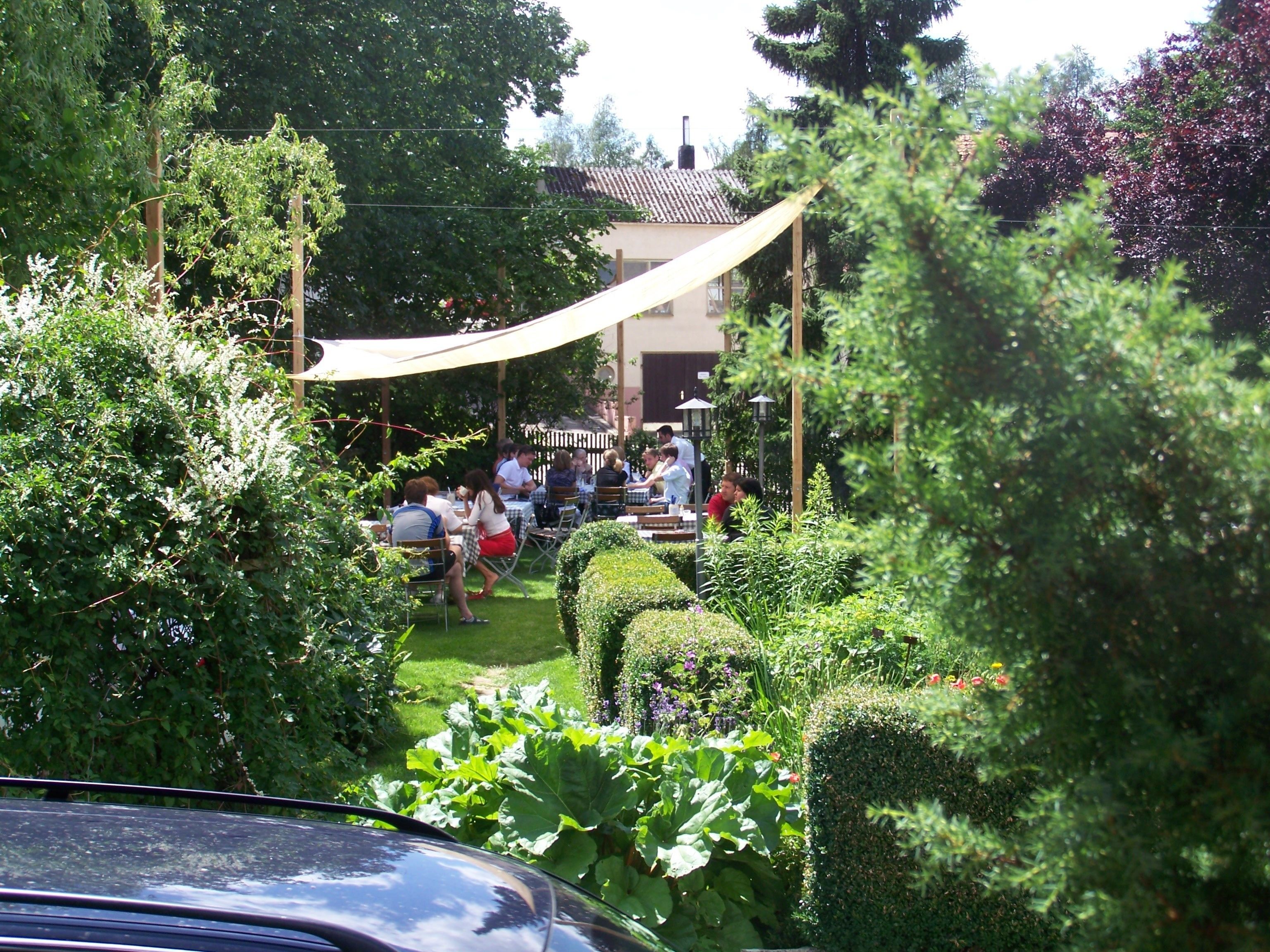 An outdoor seating area with tables and chairs, surrounded by lush greenery and a car in the foreground.