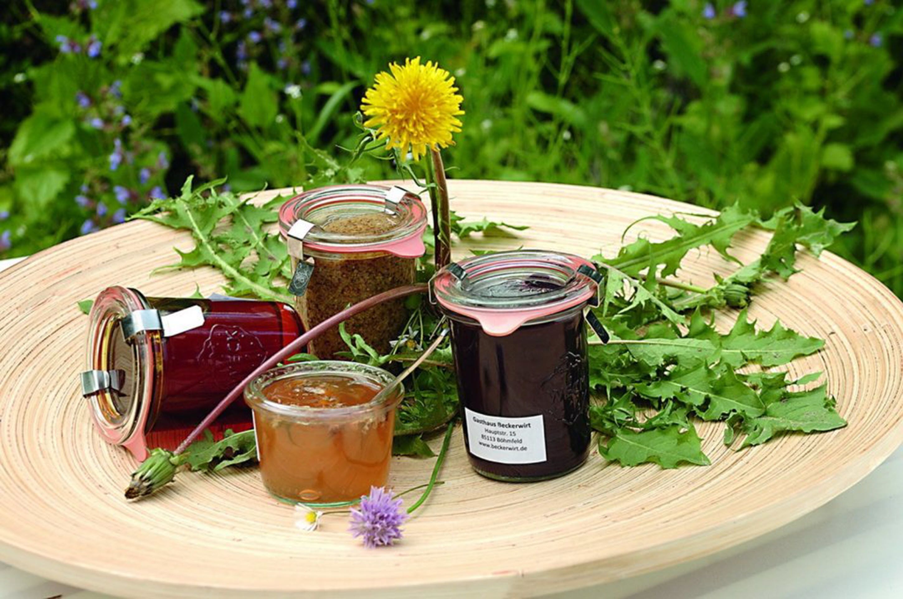 Ein Holztablett mit Gläsern voller Honig und einem Honiglöffel, umgeben von grünen Blättern und einer gelben Blüte.