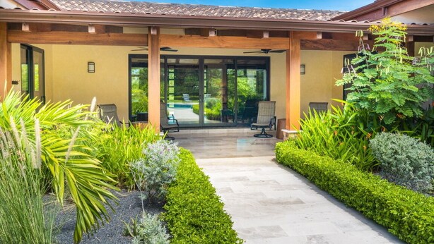 A covered outdoor area with a pathway leading to a building entrance, surrounded by well-maintained greenery and outdoor furniture.
