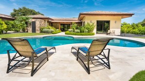A modern house with a swimming pool, two lounge chairs, and a well-maintained garden.