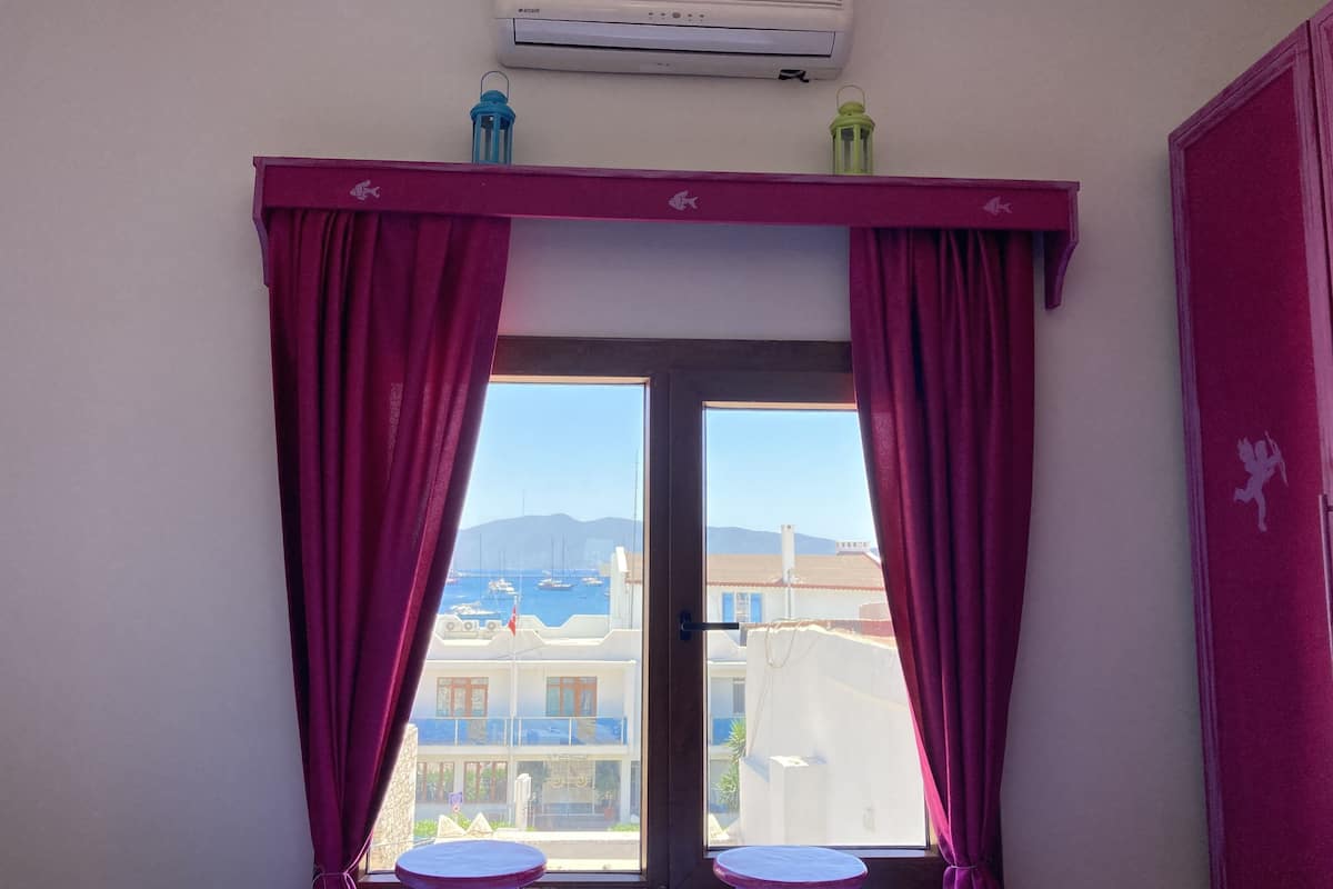 Una habitación con balcón, que incluye una ventana con cortinas moradas, una mesita y un equipo de aire acondicionado sobre la ventana.