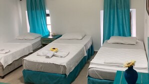 A hotel room with three single beds, each with a teal bed skirt and white linens. There is a small table with a vase of flowers, a wall-mounted air conditioner, and a window with teal curtains. - Can Hotel (Bodrum)