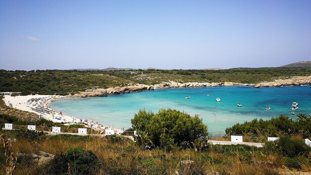Een kuststrand met helder turquoise water, een zandstrand en een achtergrond van groene heuvels.
