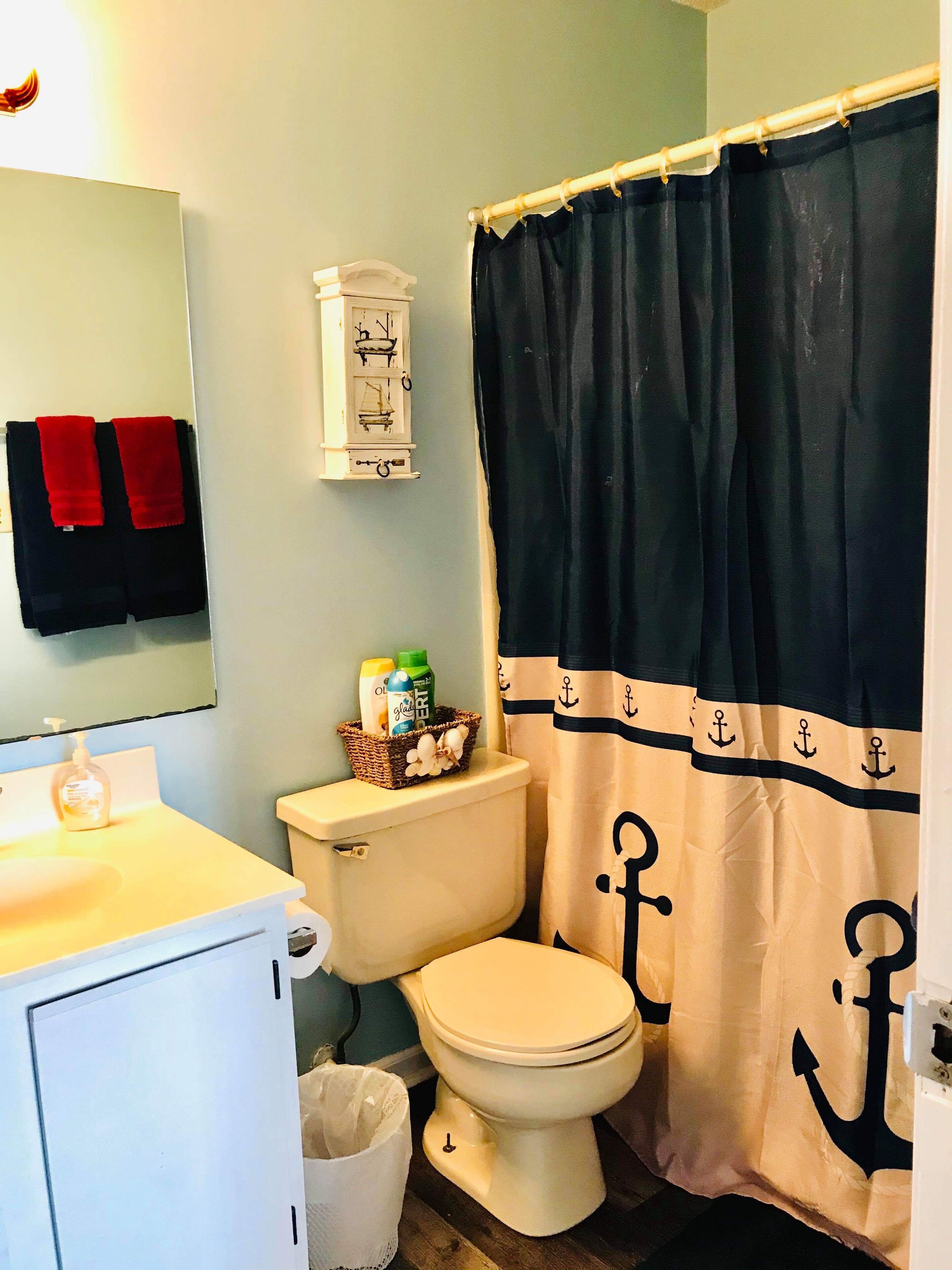 Un baño con inodoro, lavamanos, espejo, cesta de artículos de aseo y cortina de ducha con motivos de anclas.