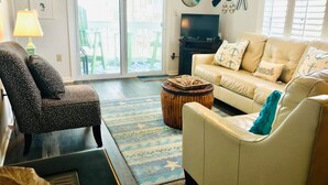 Een woonkamer met een bank, fauteuil en een televisie. Er is een raam met zonwering en een deur naar een buitenruimte.