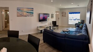 A modern living room with a large flat-screen TV, a dark blue sofa, a round dining table, and a glass-top coffee table.