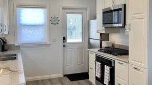 A modern kitchen with white cabinetry, a microwave, stove, and oven. There is a window with blinds and a door with a doorknob.