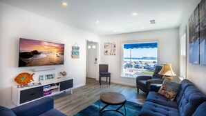 A modern living room with a large TV, comfortable seating, a view of the beach, and a window with a view of the parking lot.