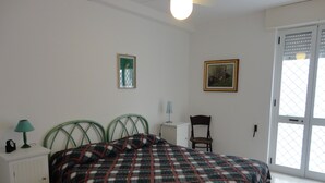 A bedroom with a bed, a nightstand with a lamp, a chair, and a door with a view. - VersoSud - Apartment Vicino Spiaggia 04 (Ugento)