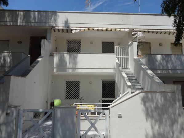 A multi-story building with a white exterior, multiple windows with metal grilles, and a staircase leading to the upper floors.