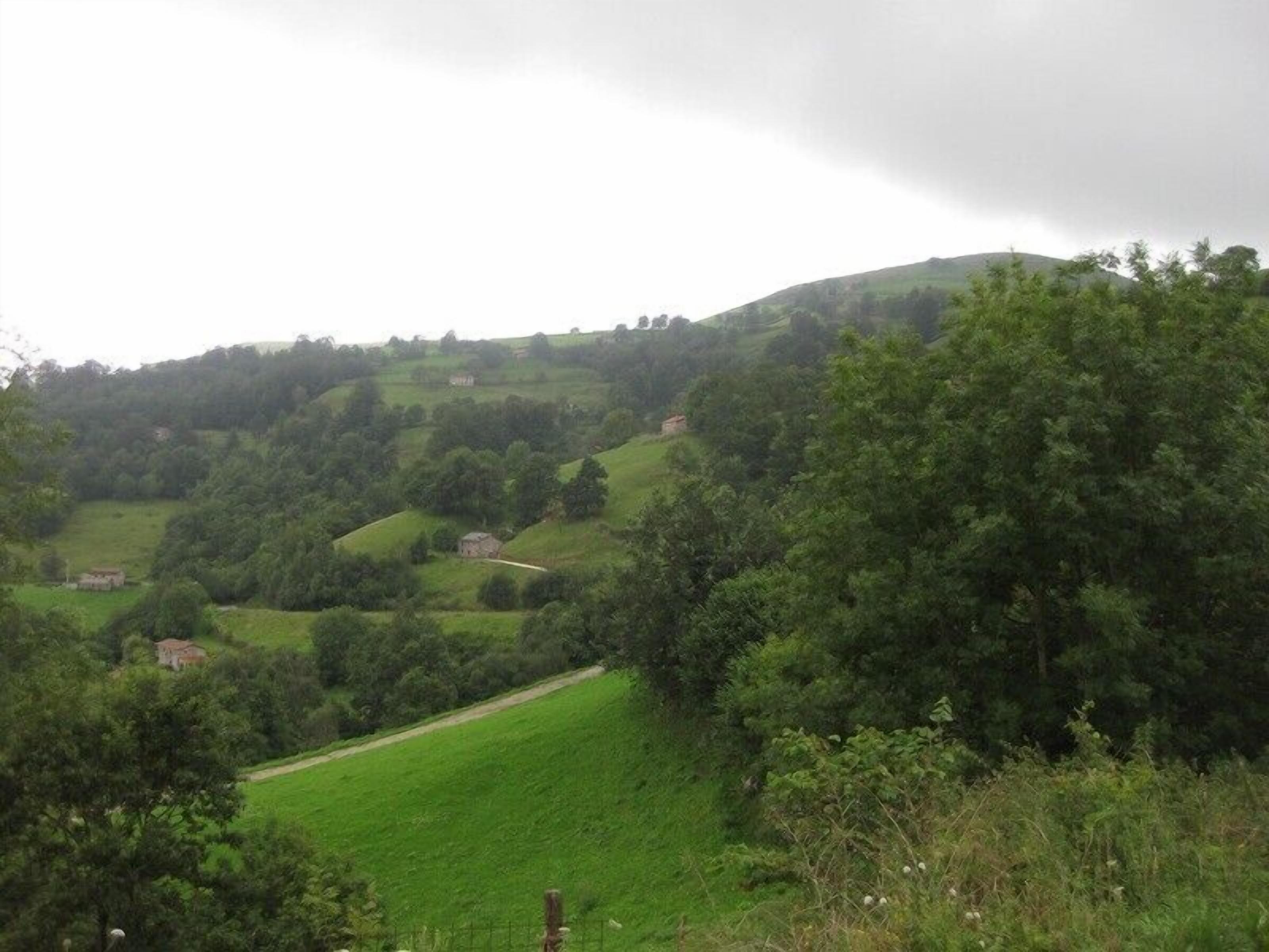 A rural landscape with rolling hills, scattered houses, and dense greenery.