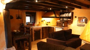 A cozy living room with a fireplace, a dining area with a brick table, and a kitchen with wooden cabinets and a hanging light fixture. - Charming cottage in the valleys pasiegos. Stunning views of the mountains. (San Roque de Riomiera)