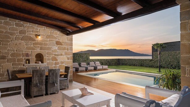 A stone-walled outdoor area with a pool, lounge chairs, and a view of mountains at sunset.