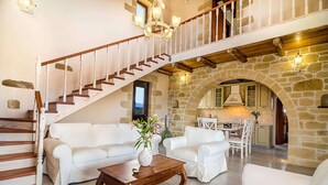A spacious living area with a staircase, white furniture, and a stone archway leading to a kitchen.