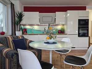 Eine moderne Küche mit weißen Schränken, ein Essbereich mit einem Glastisch und ein Sofa mit gemusterten Polstern.