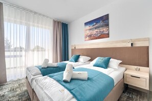 Et hotelværelse med to senge, et stort vindue med lette gardiner, et billede af en strandscene og en trægavl.