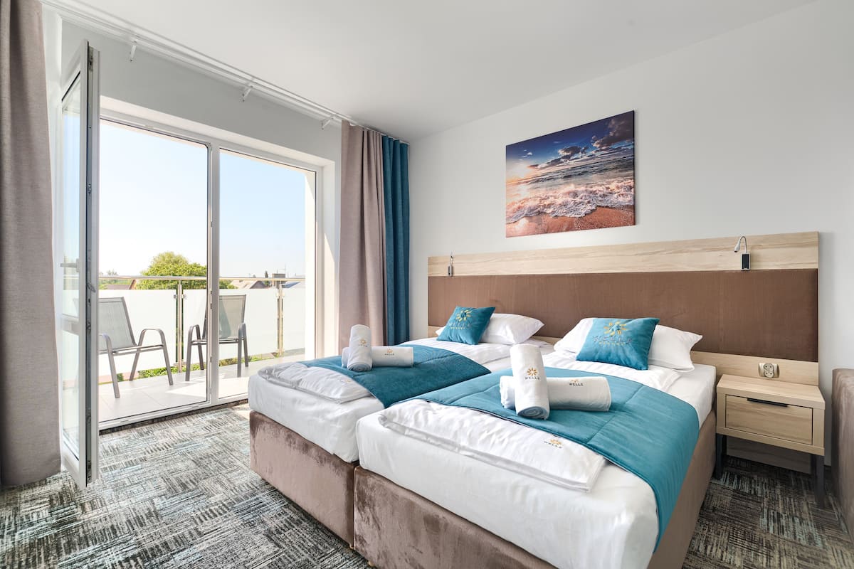 A hotel room with two beds, a large window with a view, a painting on the wall, and a small table with a lamp.