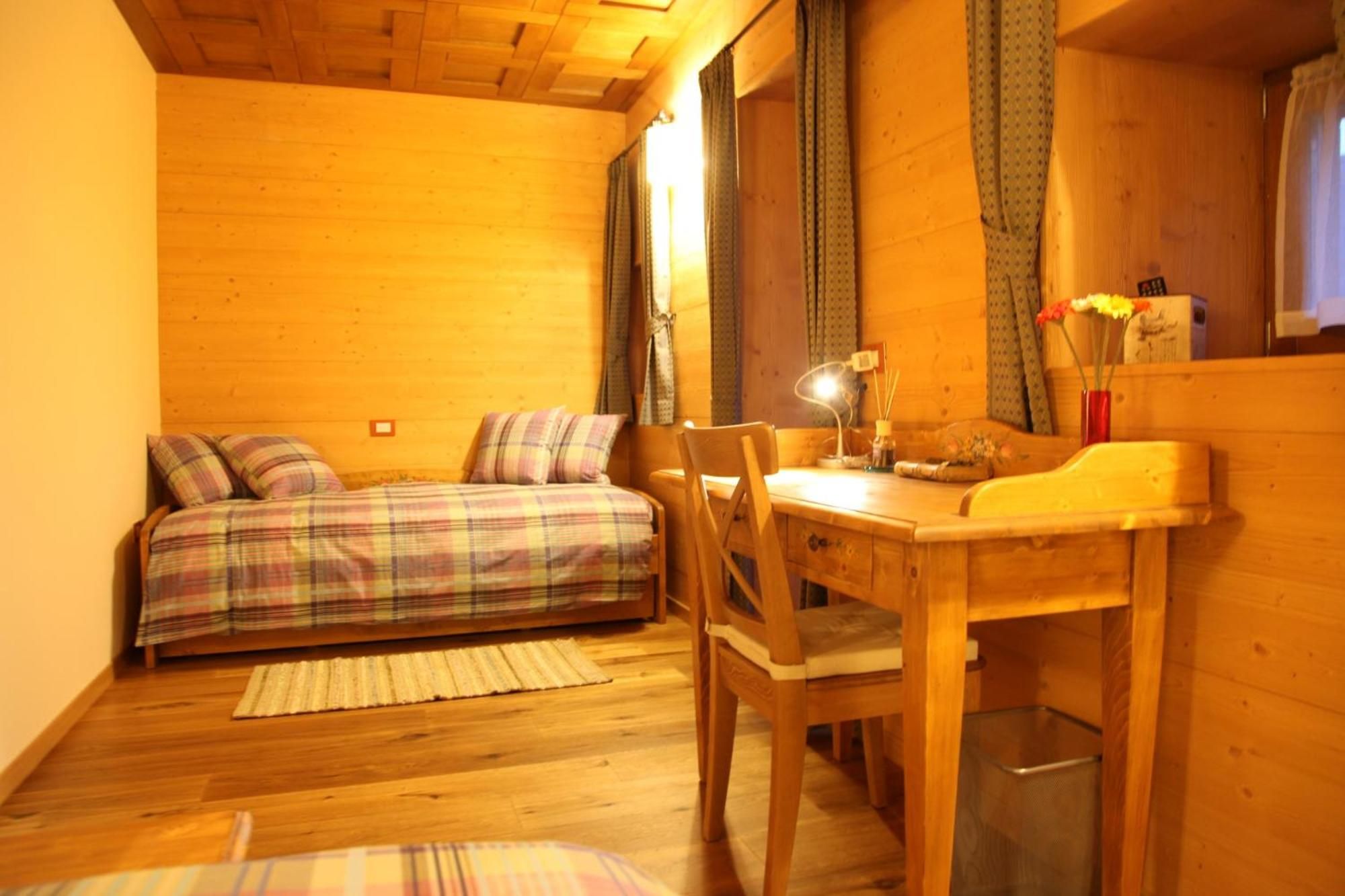 Una habitación acogedora con suelo de madera, una cama con ropa de cama escocesa, una mesa de madera con una lámpara y una ventana con cortinas.