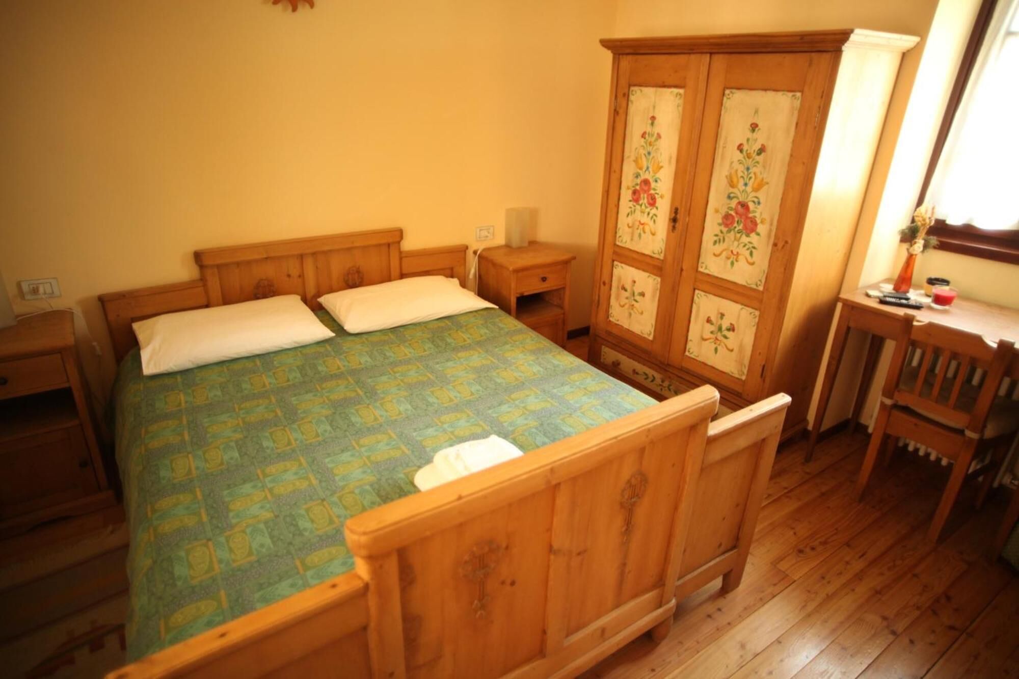 Ein hölzernes Bett mit einem gemusterten Überwurf, ein Holzschrank mit floralen Verzierungen und ein kleiner Holztisch mit einer Lampe.