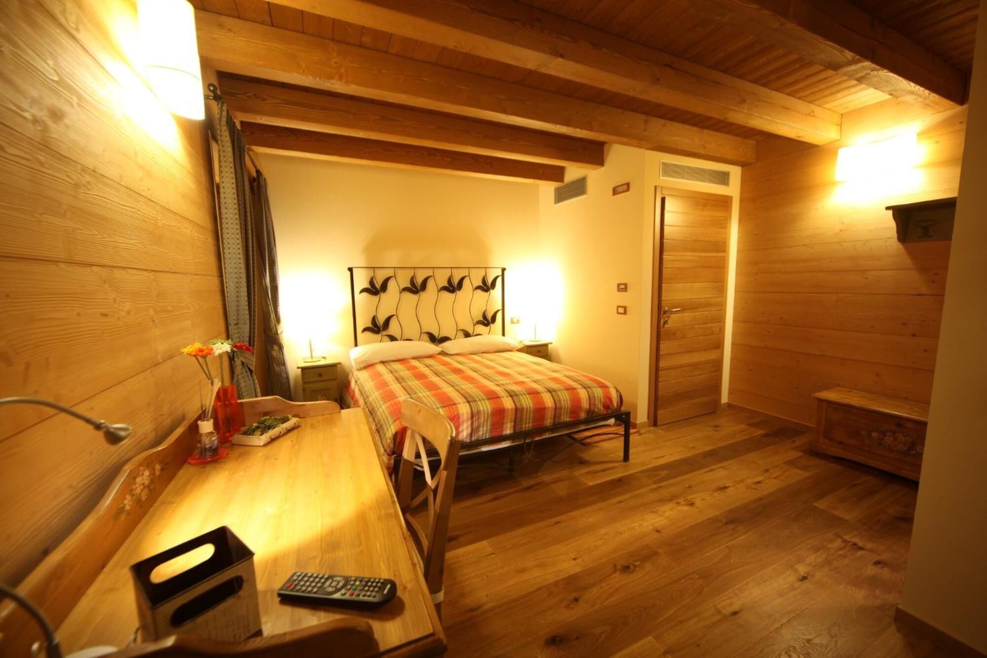 A cozy room with a bed, a wooden desk, and a chair. There is a nightstand with a lamp and a vase with flowers. The room has wooden walls and a wooden ceiling.