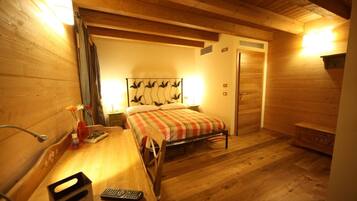 A cozy room with a bed, a wooden desk, and a chair. There is a nightstand with a lamp and a vase with flowers. The room has wooden walls and a wooden ceiling.