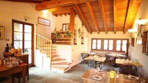 A dining area with wooden tables and chairs, a staircase leading to an upper level, and a window with blinds. - Le Case dei Baff (Ardenno)