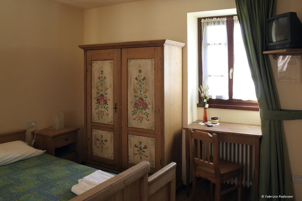 Un dormitorio con cama, un armario de madera con diseños florales, un escritorio con silla, un radiador y un televisor sobre una estantería.