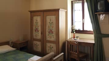 Un dormitorio con cama, un armario de madera con diseños florales, un escritorio con silla, un radiador y un televisor sobre una estantería.