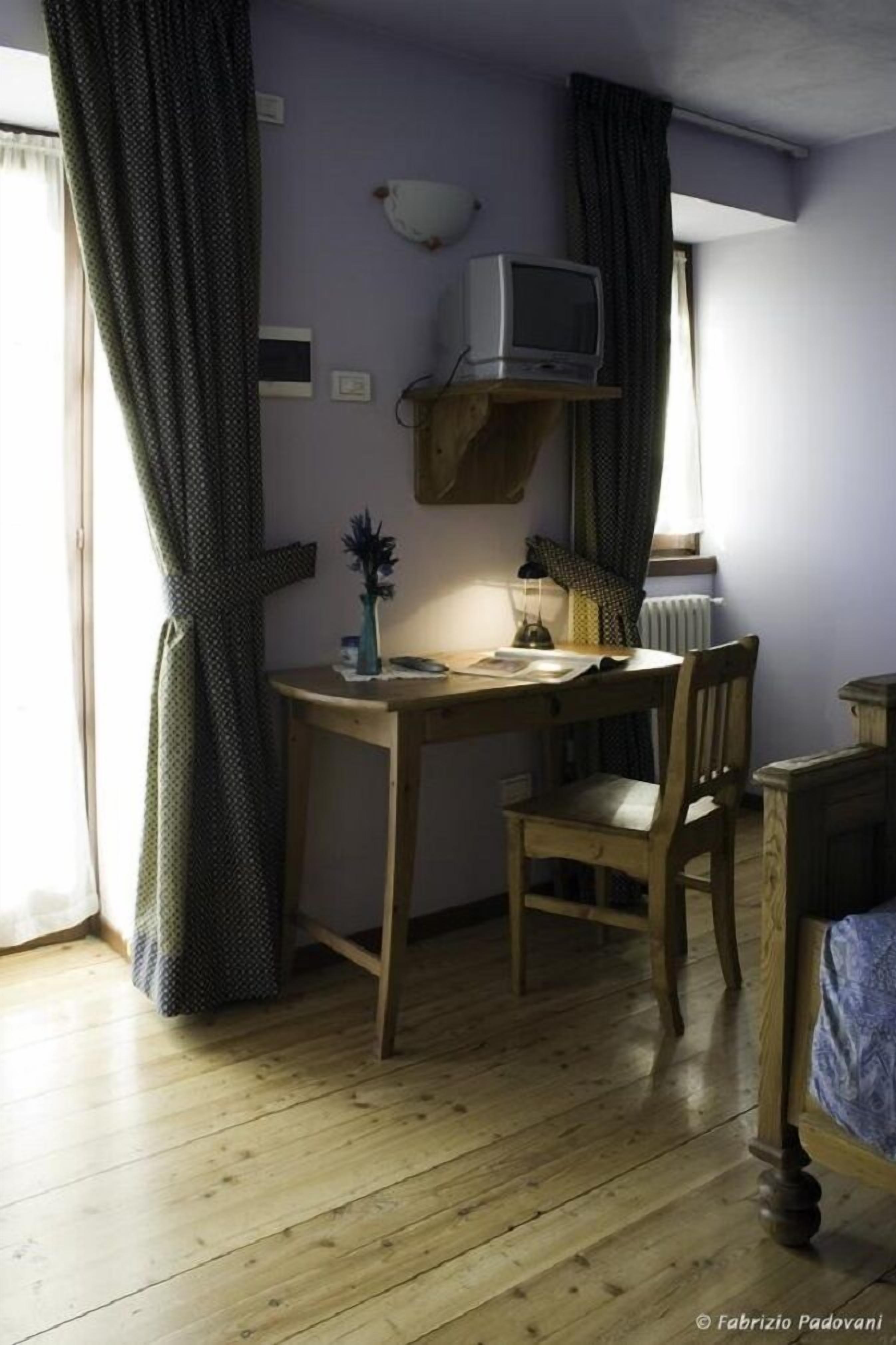 Une pièce comprenant un bureau en bois, une chaise, un lit, un téléviseur posé sur une étagère et une fenêtre avec des rideaux.